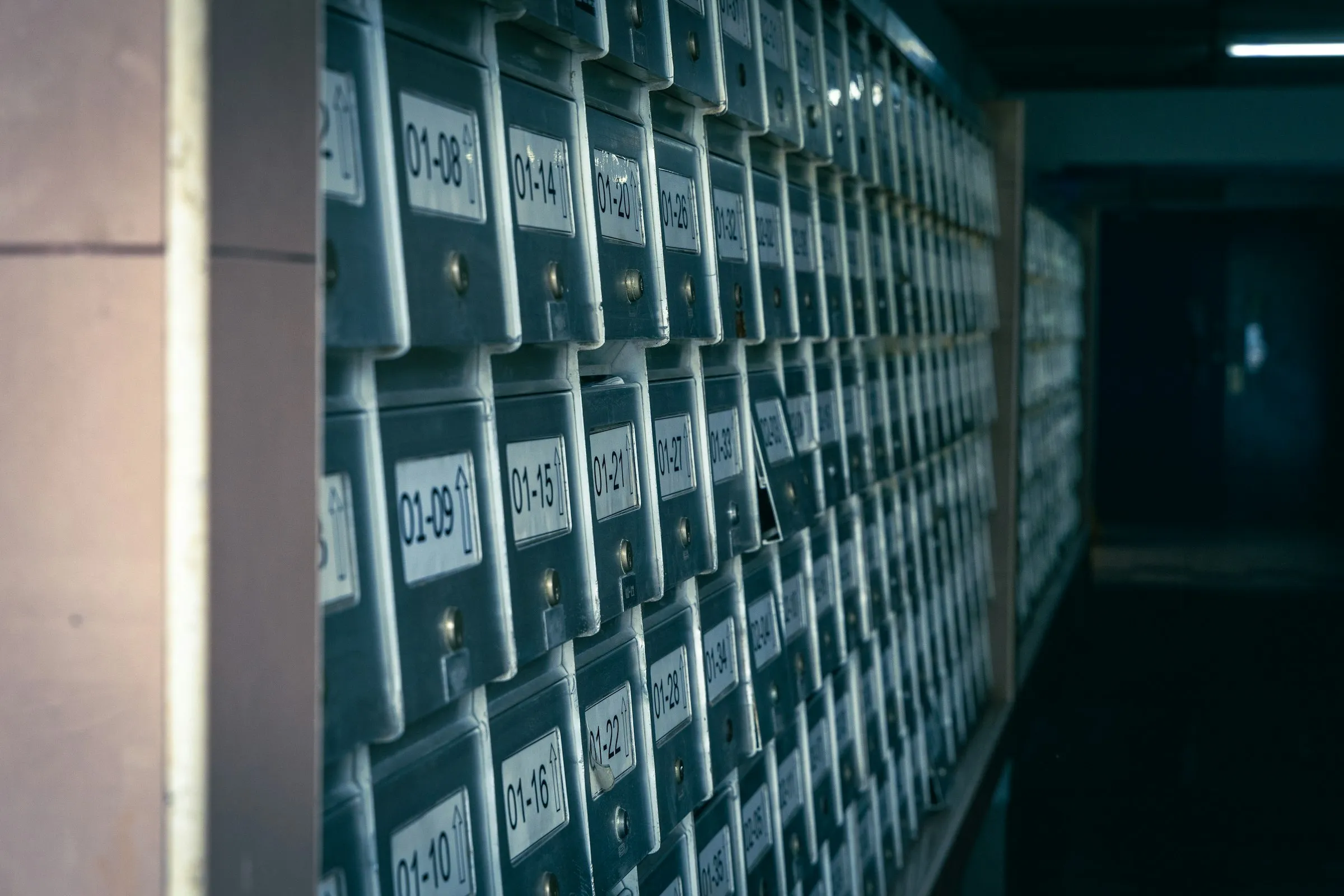 A wall of numbered metal post office boxes, each with a small keyhole and a paper label, receding into a dim corridor