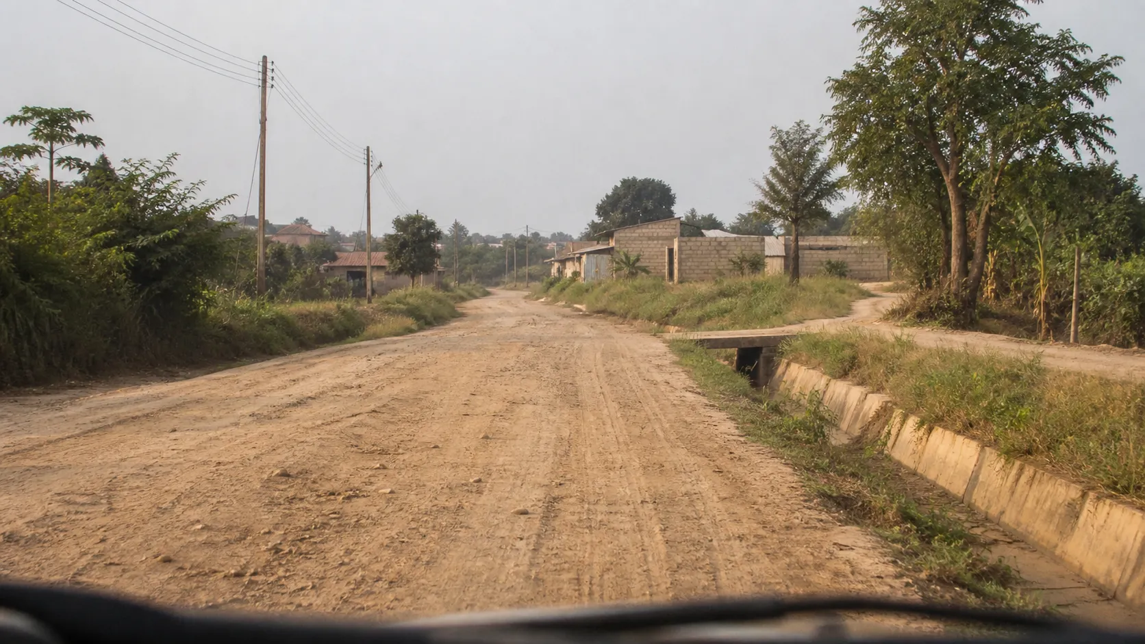 A street-level view of an unpaved rural road, including the road surface, drainage, vegetation, buildings, and utility poles